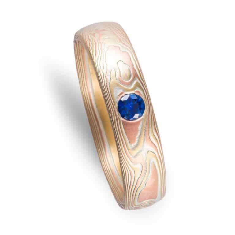 simple and lovely mokume gane wedding ring in fire palette with red and yellow golds and blue sapphire flush set in