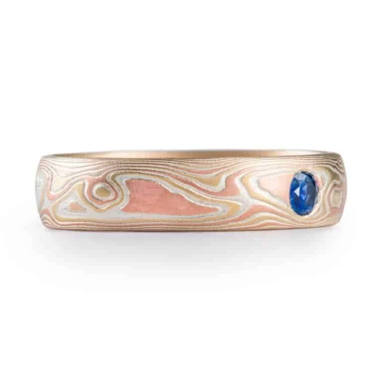 simple and lovely mokume gane wedding ring in fire palette with red and yellow golds and blue sapphire flush set in