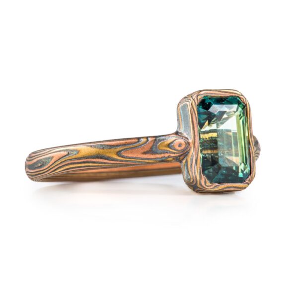 magical feeling woodgrain patterned narrow ring with a single green rectangular stone, held in a mokume patterned bezel - a narrow strip of the same material as the main ring wrapped around the perimeter of the stone