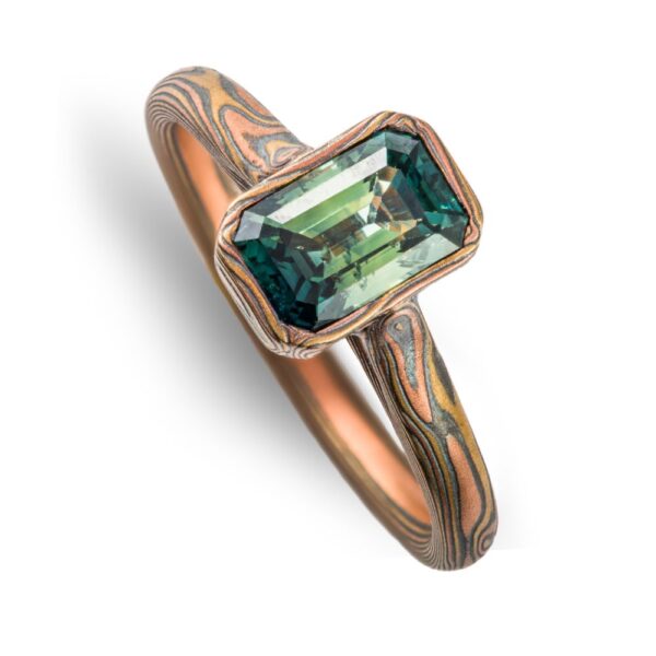 magical feeling woodgrain patterned narrow ring with a single green rectangular stone, held in a mokume patterned bezel - a narrow strip of the same material as the main ring wrapped around the perimeter of the stone