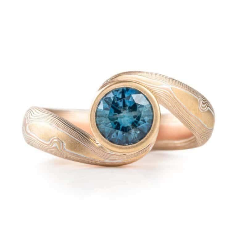 teal blue sapphire set in volume woodgrain that is a yellow gold and red gold palette . handmade goldsmith made ring