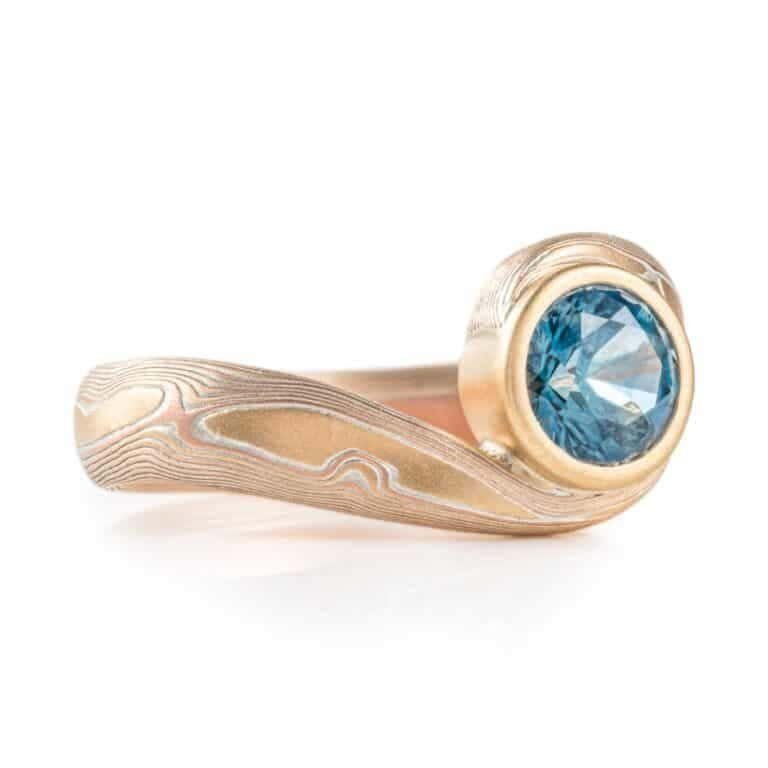 teal blue sapphire set in volume woodgrain that is a yellow gold and red gold palette . handmade goldsmith made ring