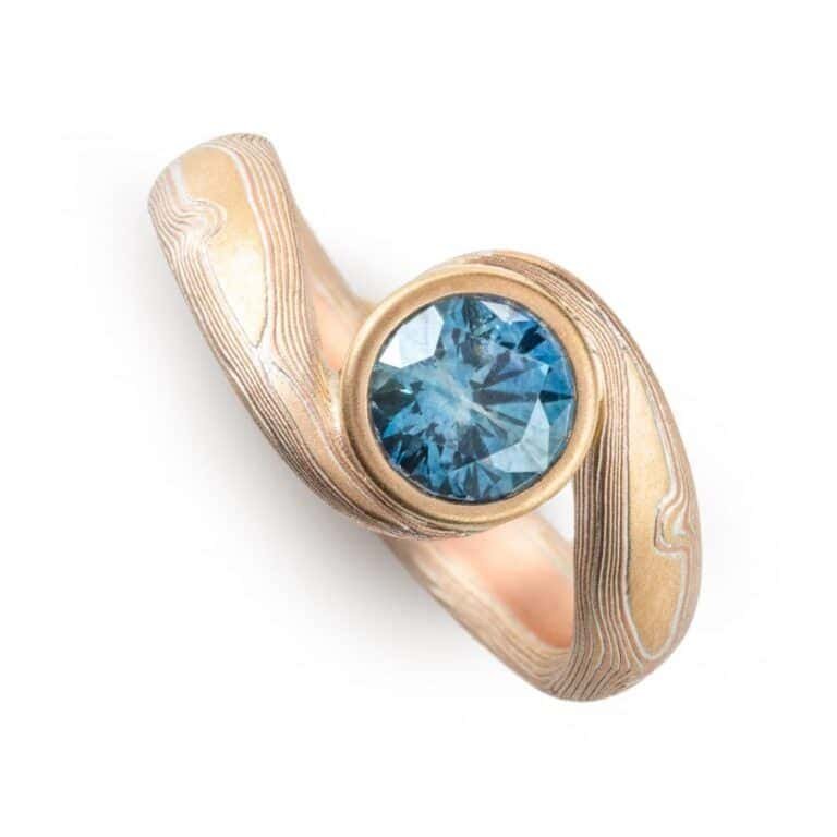 teal blue sapphire set in volume woodgrain that is a yellow gold and red gold palette . handmade goldsmith made ring