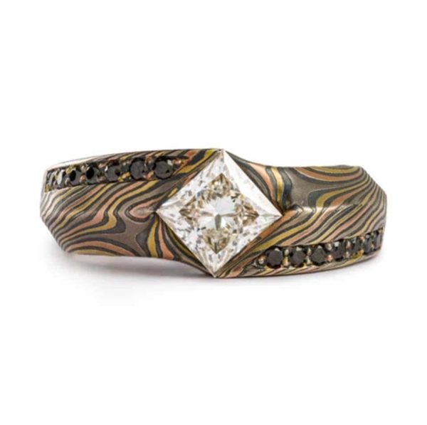 This is a custom made Mokume Gane ring, made by arn krebs, featuring a unique carved angled profile and a modified cathedral style setting holding the large white center diamond. The diamond is square and set diagonally to the band, and is flanked on each side by seven small bead set black diamonds running parallel to the band. It's made in our Twist pattern and an upgraded 18kt version of our Firestorm palette, the metals in it are red gold yellow gold palladium and silver