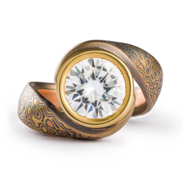 Mokume gane ring with a bypass style setting, the ring wraps itself around a large 9mm wide moissanite held in a gold bezel. The ring has a droplet pattern and the metals used are red gold yellow gold and silver.