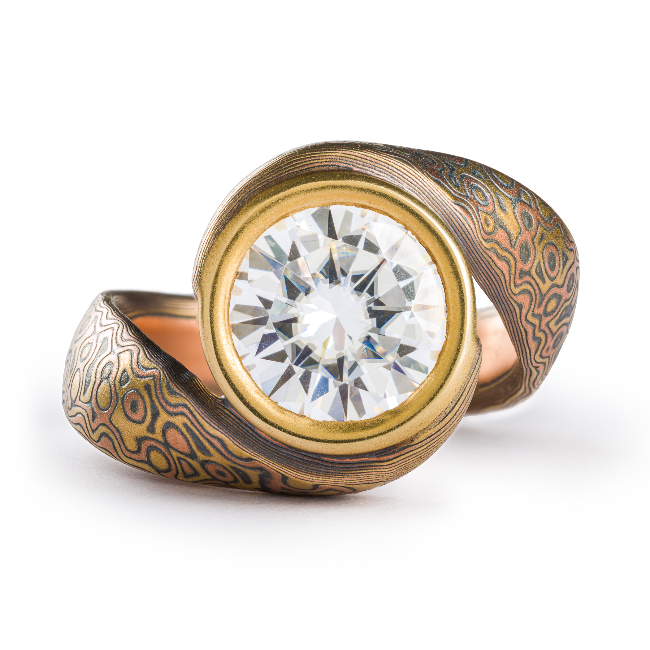 Mokume gane ring with a bypass style setting, the ring wraps itself around a large 9mm wide moissanite held in a gold bezel. The ring has a droplet pattern and the metals used are red gold yellow gold and silver.