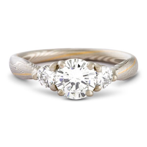 Engagement ring with large center stone flanked by a smaller diamond on either side, set in prongs. The band is made of mokume gane in shades of silver and gray with one stripe of yellow gold.