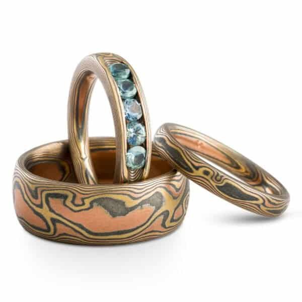 Set of three mokume gane rings, patterned to look like a warm toned wood. One wider band, one narrower, and one narrower with light blue stones set into it in a line.
