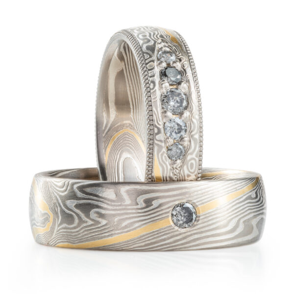 Matching set of mokume gane rings, one with a row of salt and pepper diamonds bead set into the band, with textured rails going around each edge. Both rings are made with silver and light gray metals, with one stripe of yellow gold.