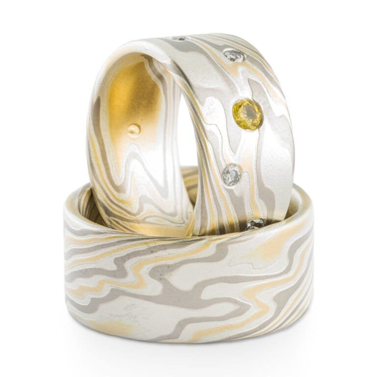 Set of mokume gane rings, both wide with flat profiles. Made with yellow gold, palladium and silver, in matching pattern. One ring has small flush set yellow and white stones.