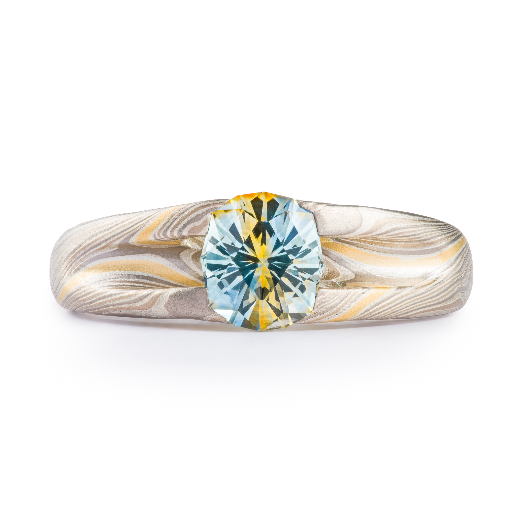 Mokume gane patterned ring with large multicolored sapphire held in prongs carved from the body of the ring. The stone is a light blue color, with a gold stripe running through the center, echoing the gold stripe running throughout the rest of the band.