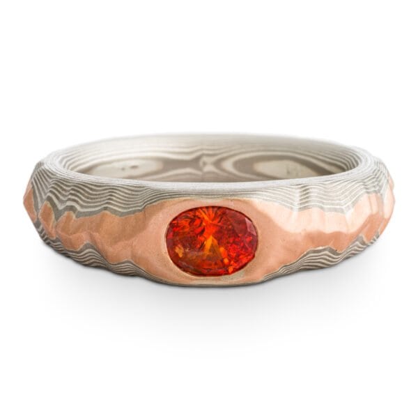 Mokume gane ring with oval deep orange sapphire, the ring is patterned to look like a mountain range, the main ring is gray and silver, topped with red gold.