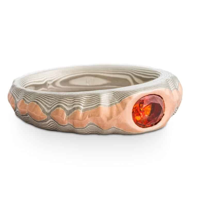 Mokume gane ring with oval deep orange sapphire, the ring is patterned to look like a mountain range, the main ring is gray and silver, topped with red gold.