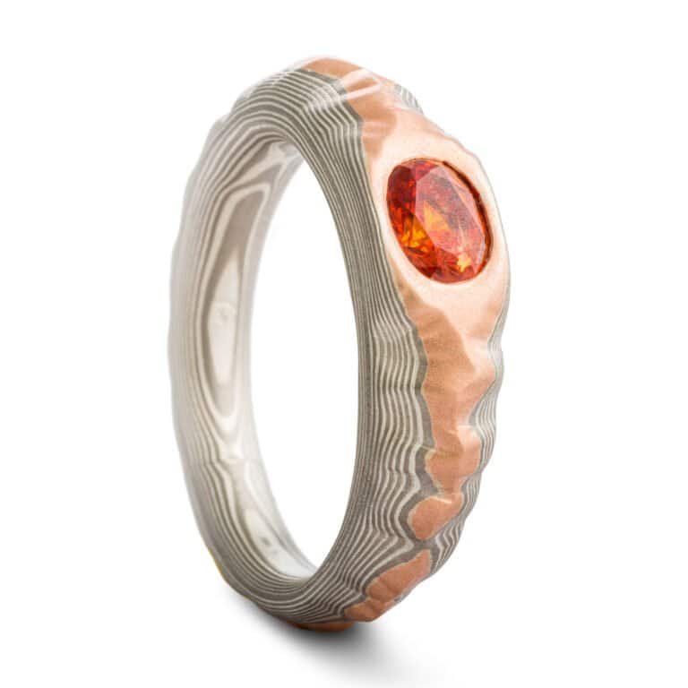 Mokume gane ring with oval deep orange sapphire, the ring is patterned to look like a mountain range, the main ring is gray and silver, topped with red gold.