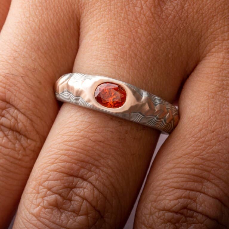 Mokume gane ring with oval deep orange sapphire, the ring is patterned to look like a mountain range, the main ring is gray and silver, topped with red gold.