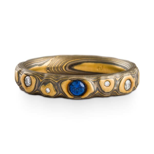Narrow mokume gane ring made with yellow gold, palladium and oxidized silver, in a three dimensional pattern made to resemble rocks. The ring has small flush set stones, one larger blue one at the center, and smaller white stones placed organically throughout.