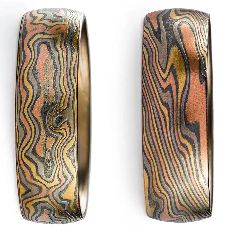 two mokume gane rings side by side in the same metal combination, the one on the left is shown in woodgrain pattern, the one on the right is shown in twist pattern to highlight the differences in pattern between the two, woodgrain follows the direction of the band, twist runs diagonally across the band