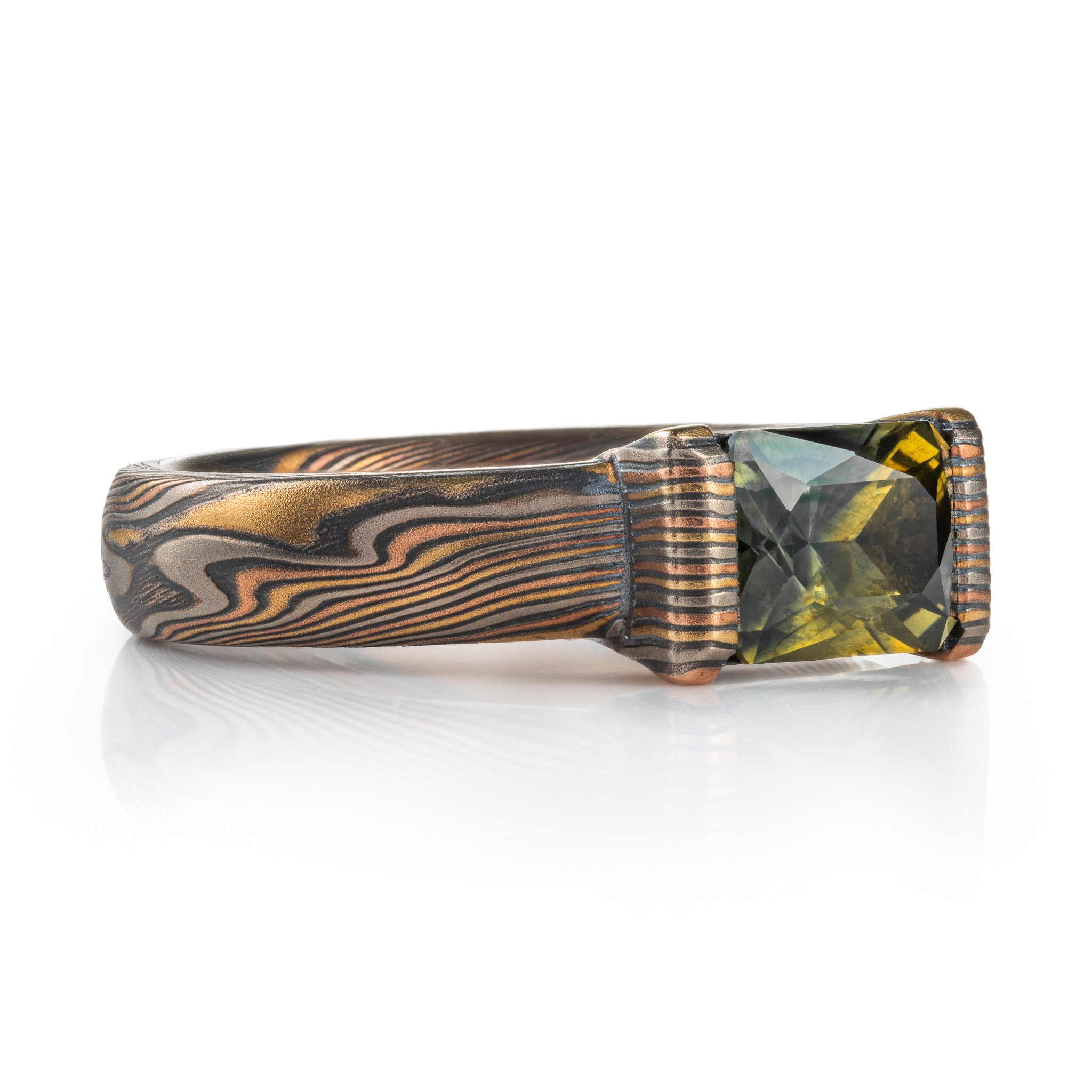 whimsical and unique engagement style mokume gane ring, shown in Firestorm palette with matching mokume patterned bezel, holding a rectangular mossy green sapphire