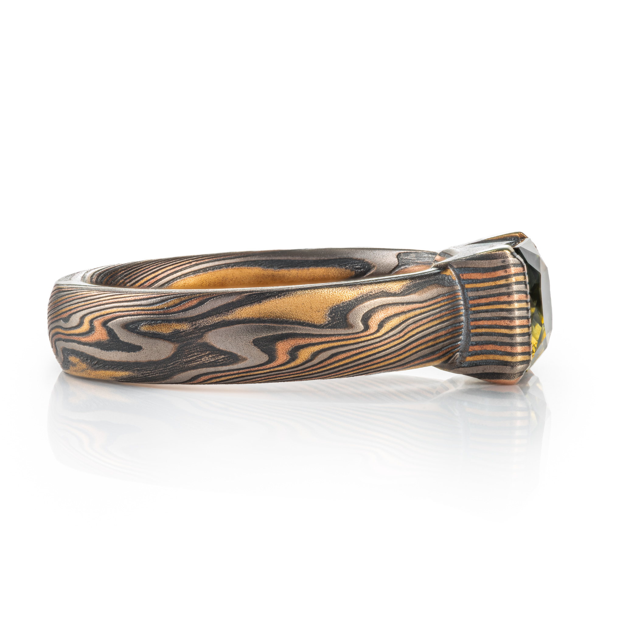 whimsical and unique engagement style mokume gane ring, shown in Firestorm palette with matching mokume patterned bezel, holding a rectangular mossy green sapphire