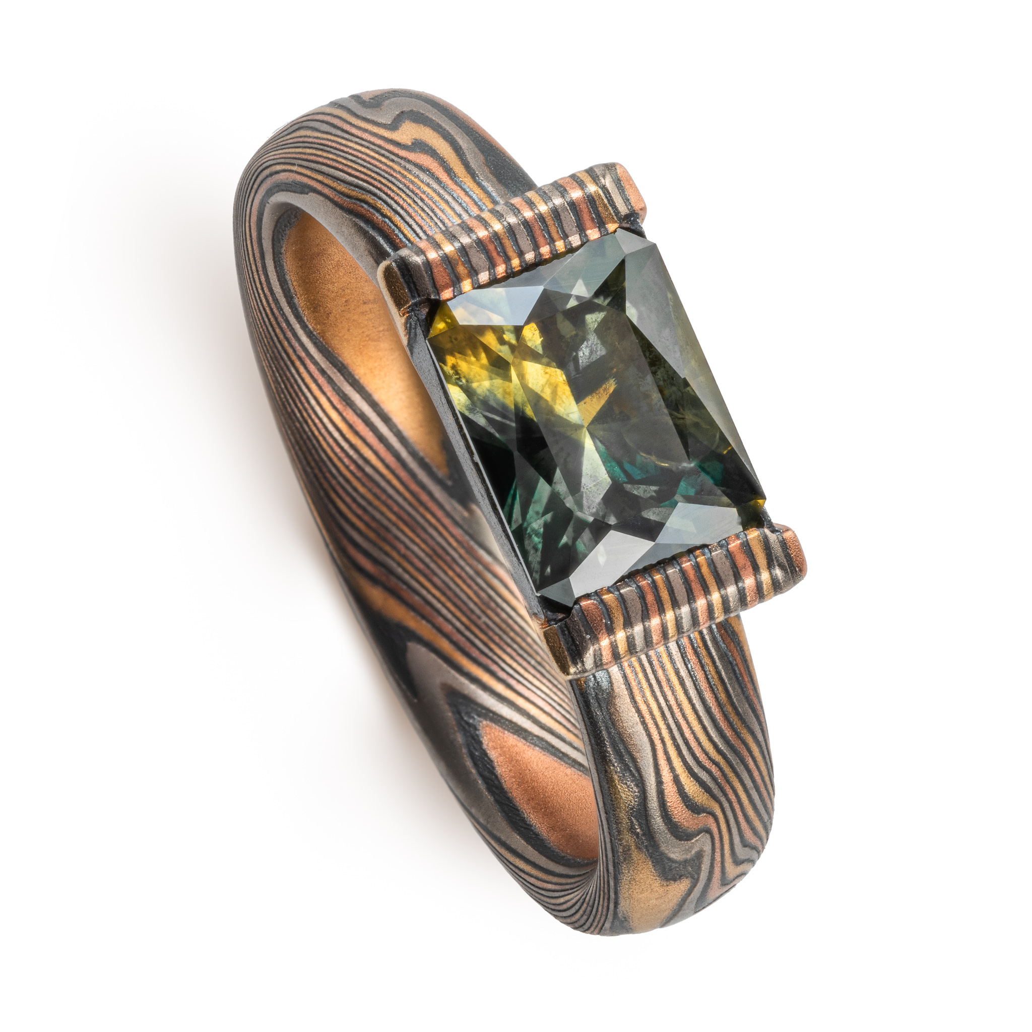 whimsical and unique engagement style mokume gane ring, shown in Firestorm palette with matching mokume patterned bezel, holding a rectangular mossy green sapphire