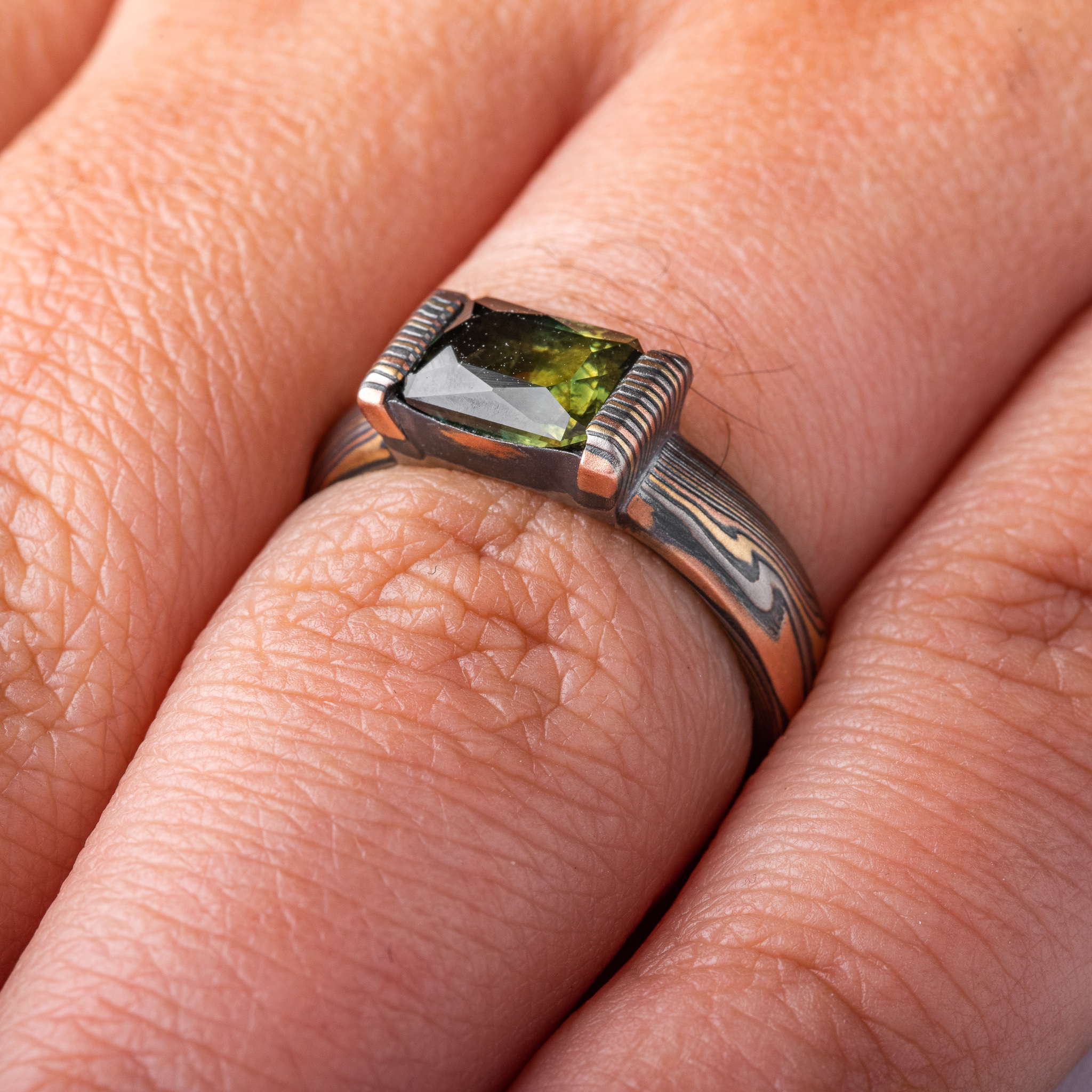 whimsical and unique engagement style mokume gane ring, shown in Firestorm palette with matching mokume patterned bezel, holding a rectangular mossy green sapphire