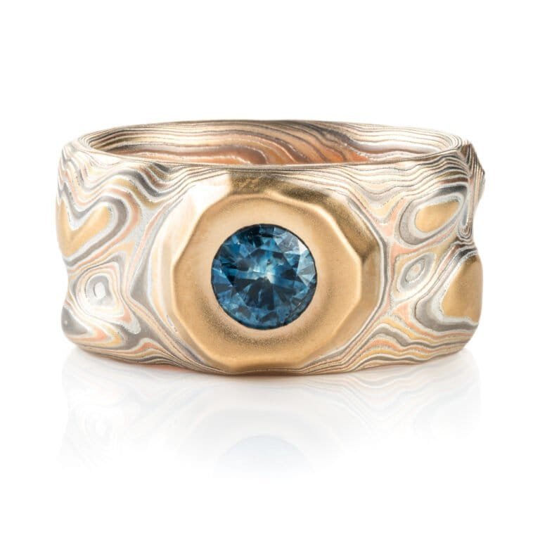 wide man ring in mokume of yellow gold, red gold, silver, and palladium with a recessed blue sapphire. handmade and heirloom quality