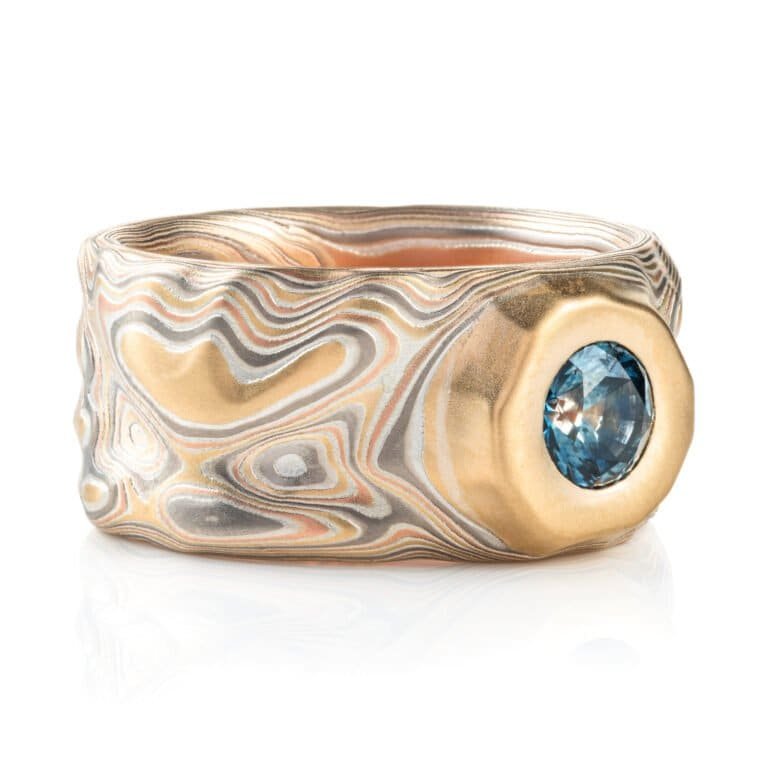 wide man ring in mokume of yellow gold, red gold, silver, and palladium with a recessed blue sapphire. handmade and heirloom quality