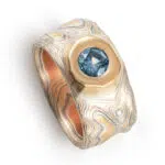 wide man ring in mokume of yellow gold, red gold, silver, and palladium with a recessed blue sapphire. handmade and heirloom quality