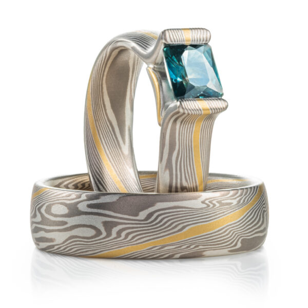 pair of mokume gane rings, one stood up vertical, the other laying flat, the upright ring has a rectangular blue stone set into it.