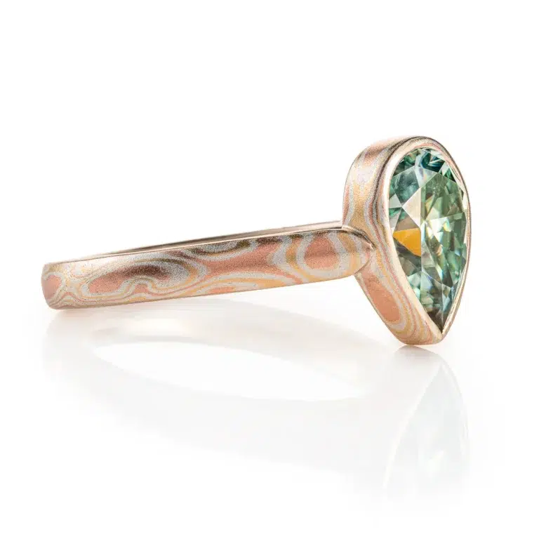 mokume gane ring with green moissanite in a pear shape, the stone is held in a mokume bezel, bezel and ring are patterned in woodgrain, with red gold yellow gold and silver