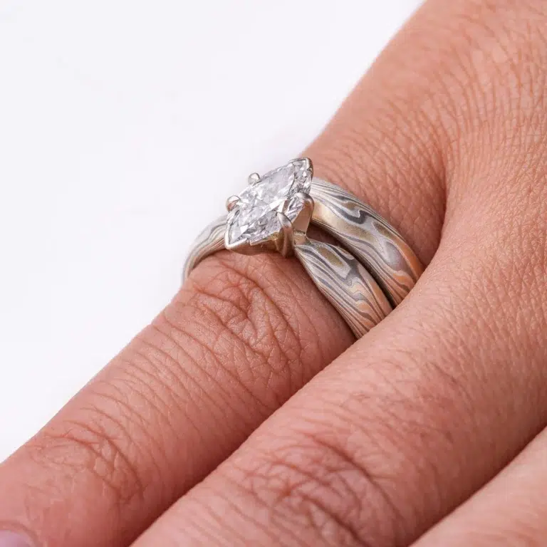 pair of stacked rings patterned in mokume twist style, the ring on top has a single marquise shaped diamond, that is wider than the band it is on and hangs over the front of the band underneath it