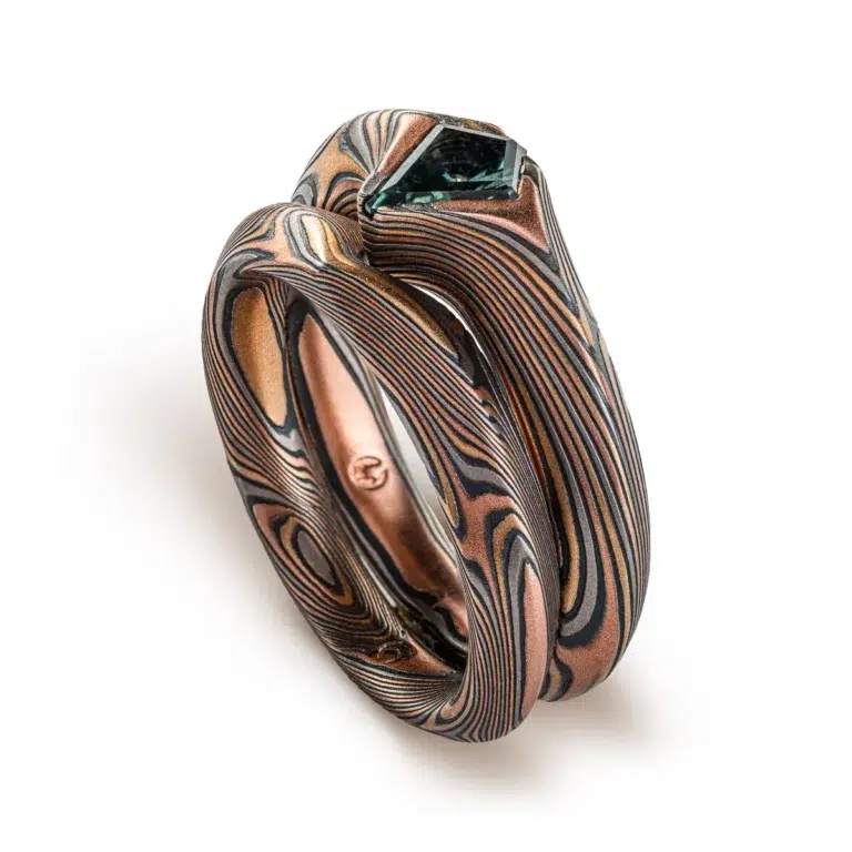 pair of mokume gane rings nested together, stacked one on top of the other, one with a kite shaped green stone set into it