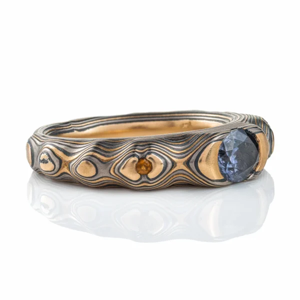 Elegant mokume gane ring with a cathedral style setting, three stones, one larger oval shaped stone at the center, and slightly smaller round stones on either side. Ring is patterned in style made to resemble mountainous terrain.