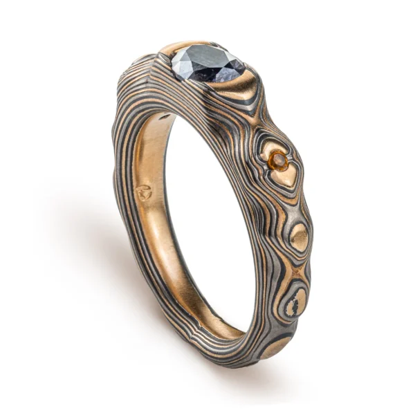 Elegant mokume gane ring with a cathedral style setting, three stones, one larger oval shaped stone at the center, and slightly smaller round stones on either side. Ring is patterned in style made to resemble mountainous terrain.