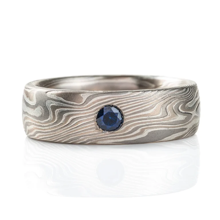 This domed band is patterned in the mokume gane twist style, in a silvery white metal combination, with one blue round stone set flush into the band.