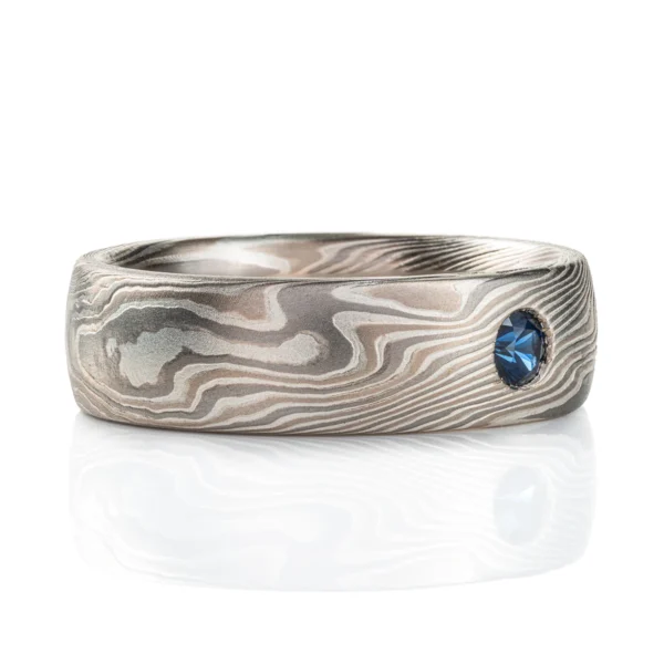 This domed band is patterned in the mokume gane twist style, in a silvery white metal combination, with one blue round stone set flush into the band.