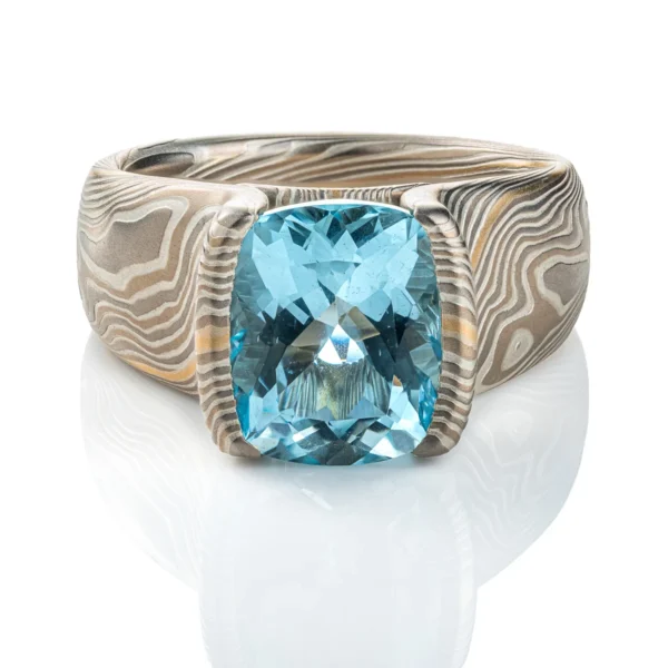 Large statement ring made in twisting mokume gane pattern. Ring is overall light and dark gray with one streak of yellow gold. The ring features a very large blue/green stone, slightly rounded rectangle shape.