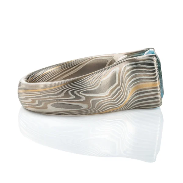 Large statement ring made in twisting mokume gane pattern. Ring is overall light and dark gray with one streak of yellow gold. The ring features a very large blue/green stone, slightly rounded rectangle shape.