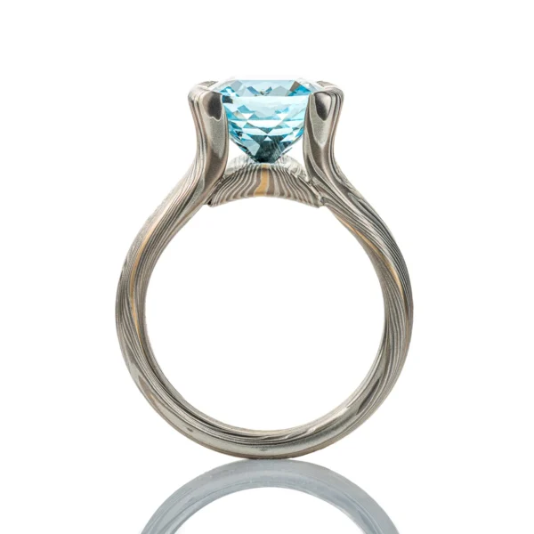 Large statement ring made in twisting mokume gane pattern. Ring is overall light and dark gray with one streak of yellow gold. The ring features a very large blue/green stone, slightly rounded rectangle shape.