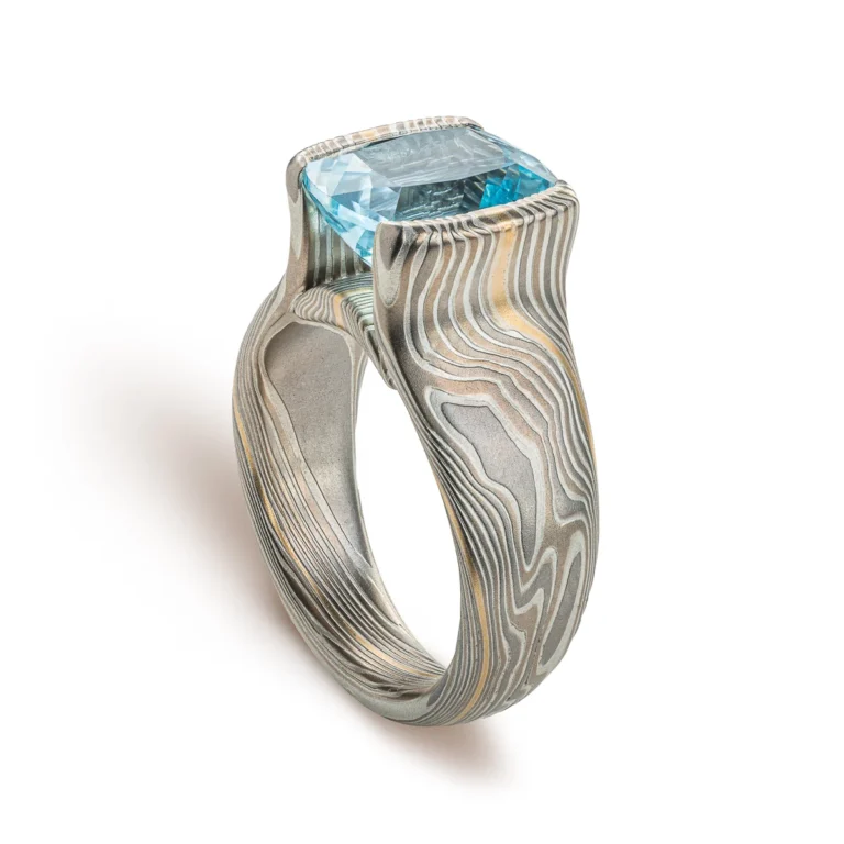 Large statement ring made in twisting mokume gane pattern. Ring is overall light and dark gray with one streak of yellow gold. The ring features a very large blue/green stone, slightly rounded rectangle shape.