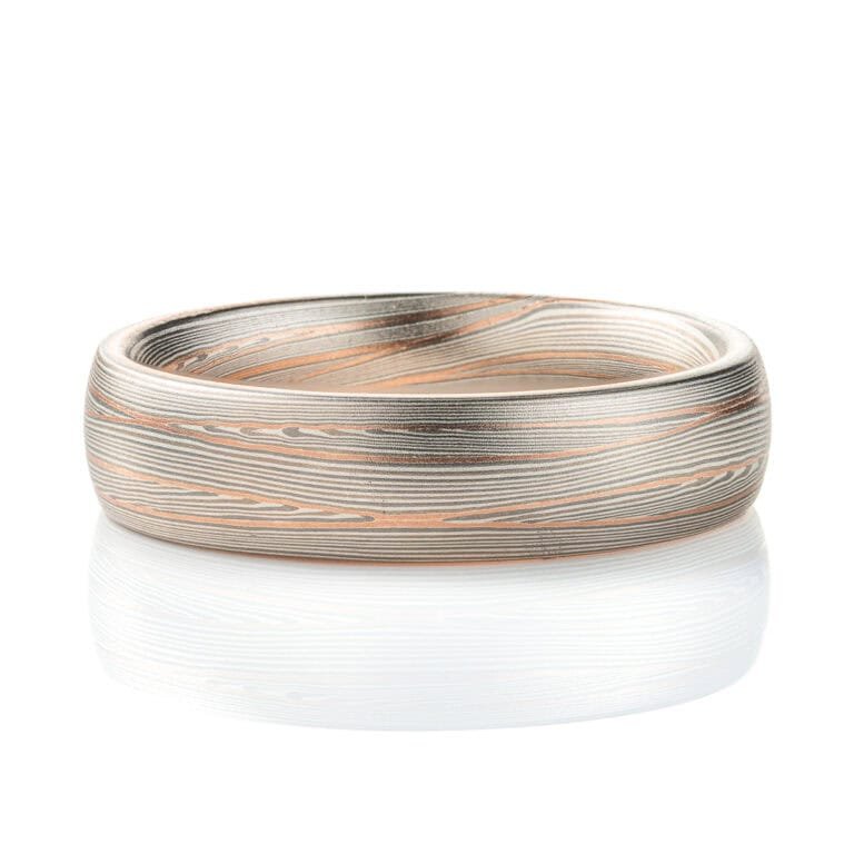 Domed band patterned in mokume style with long thin lines flowing around the ring, made of layers of light and darker gray silver metal, with two layers of red gold added.