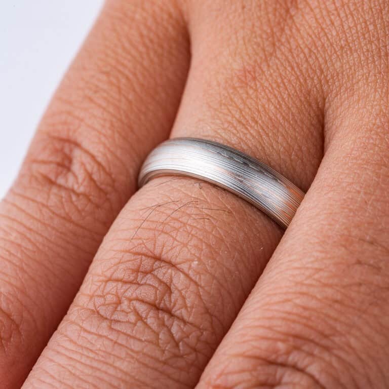 Domed band patterned in mokume style with long thin lines flowing around the ring, made of layers of light and darker gray silver metal, with two layers of red gold added.