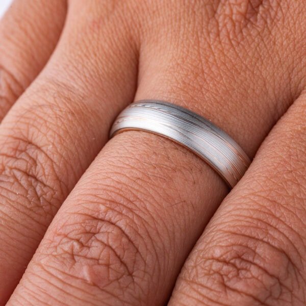 Domed band patterned in mokume style with long thin lines flowing around the ring, made of layers of light and darker gray silver metal, with two layers of red gold added.