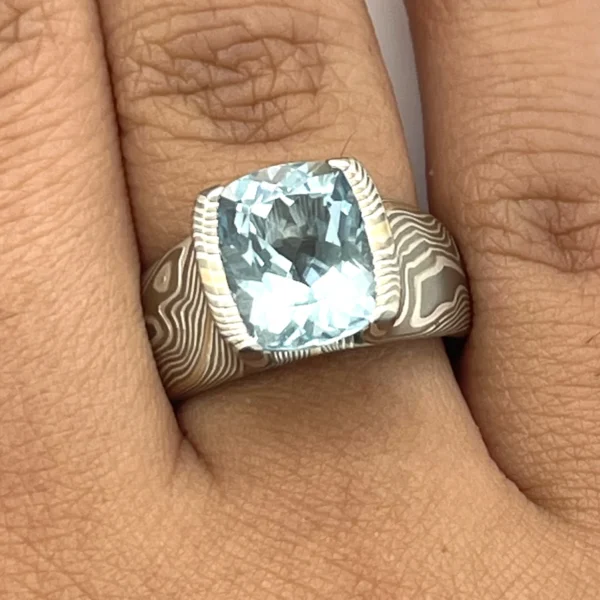 Large statement ring made in twisting mokume gane pattern. Ring is overall light and dark gray with one streak of yellow gold. The ring features a very large blue/green stone, slightly rounded rectangle shape.