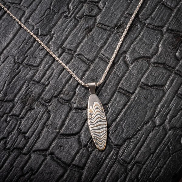 Slim oval shaped mokume gane patterned pendant, made with layers of yellow gold, palladium and silver, on a silver chain. .