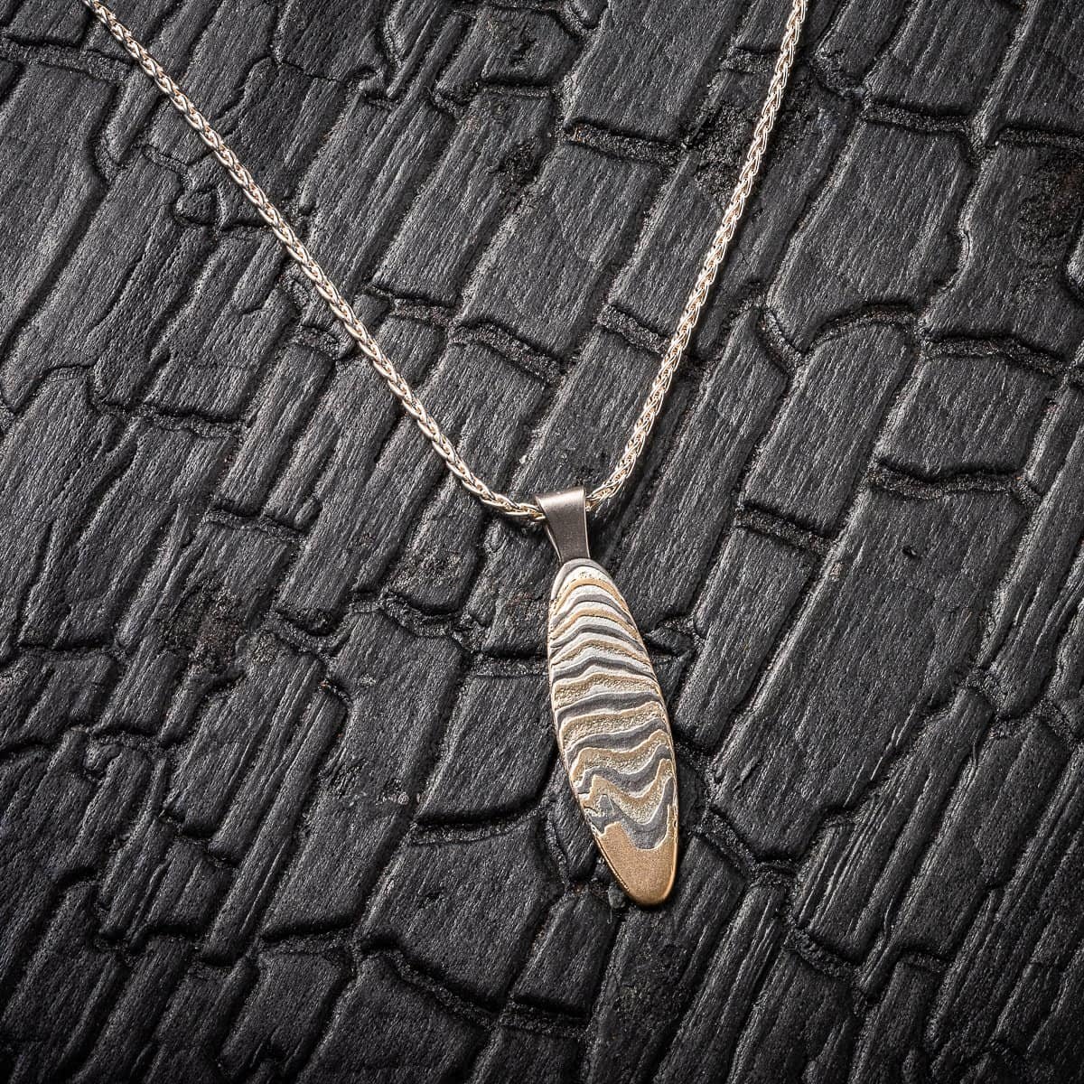 Slim oval shaped mokume gane patterned pendant, made with layers of yellow gold, palladium and silver, on a silver chain. .