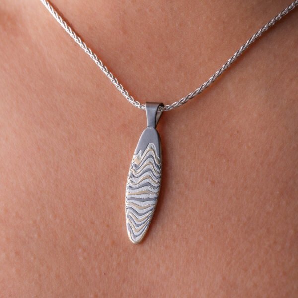 Slim oval shaped mokume gane patterned pendant, made with layers of yellow gold, palladium and silver, on a silver chain. .