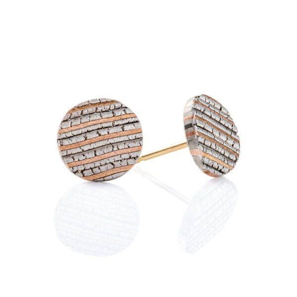 Small flat round stud earrings made with mokume gane, layered stripes of red gold, palladium and silver, with a crackly texture that resembles rock.