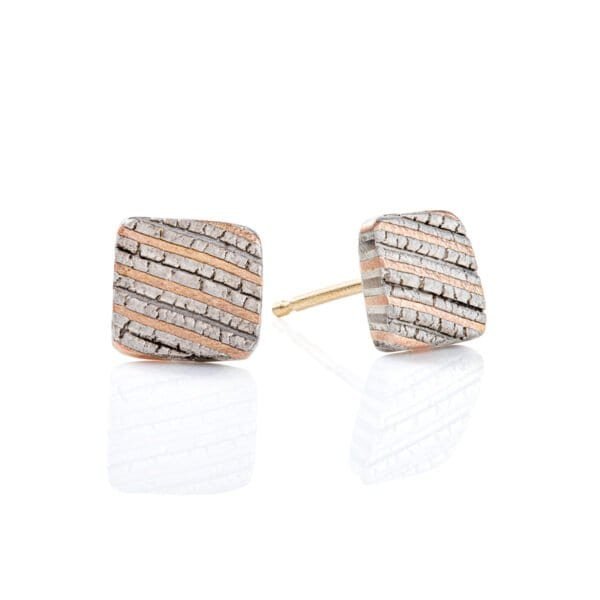 Small flat square stud earrings made with mokume gane, layered stripes of red gold, palladium and silver, with a crackly texture that resembles rock.