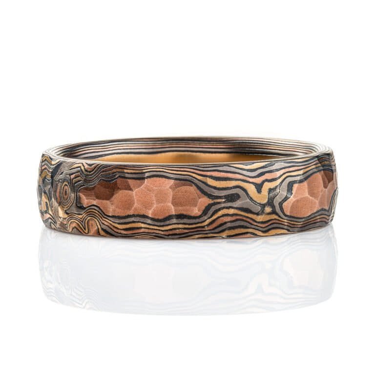 Mokume gane ring with a domed profile and textured surface all over, made with layers of red gold, yellow gold, palladium and oxidized silver (black), made in a twisting pattern that moves diagonally across the ring.